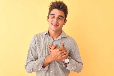Genç yakışıklı adam kapalı gözleri ve yüzünde minnettar jest ile göğüs üzerinde gülümseyerek izole sarı arka plan üzerinde duran rahat gömlek giyiyor. Sağlık kavramı.