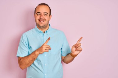 Mavi gömlekli genç adam izole edilmiş pembe arka planda duruyor gülümsüyor ve kameraya bakıyor iki eliyle ve parmaklarıyla kenarı işaret ediyor..