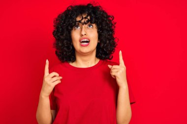 Kıvırcık saçlı genç Arap kadın izole edilmiş kırmızı arka plan üzerinde gündelik tişört giyiyor. Şaşırmış ve şaşırmış bir şekilde yukarı bakıyor ve parmaklarıyla işaret ediyor ve kollarını kaldırıyor..