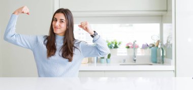 Geniş açı resim güzel genç kadın evde beyaz masaya gösteren kol kasları gururlu gülümseyen oturuyordu. Fitness kavramı.