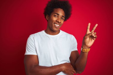 Afro saçlı, beyaz tişörtlü, izole edilmiş kırmızı arka planda duran mutlu yüzüyle kameraya bakıp zafer işareti yapan genç bir Amerikalı. İki numara..