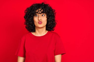 Kıvırcık saçlı genç Arap kadın izole edilmiş kırmızı arka plan üzerine günlük tişört giyip dudaklı balık suratı yapıyor, çılgınca ve komik bir jest. Komik ifade.