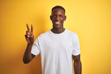 Beyaz t-shirt giyen genç Afro-Amerikalı adam izole edilmiş sarı arka planda duruyor ve iki numarayı gösterirken kendinden emin ve mutlu gülümsüyor..