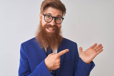 Genç, kızıl saçlı, takım elbiseli ve gözlüklü bir iş adamı beyaz arka plan üzerinde hayrete düşmüş ve kameraya gülümserken eliyle ve parmağıyla işaret ediyordu..