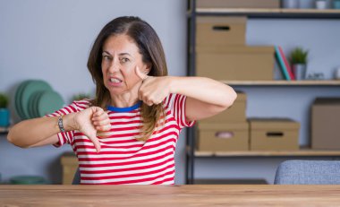 Orta yaş yaşlı kadın evde masada oturan başparmak yukarı ve aşağı yapıyor, anlaşmazlık ve anlaşma ifade. Çılgın çatışma