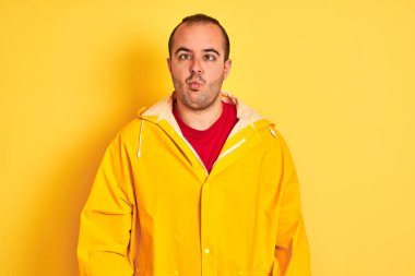 İzole edilmiş sarı arka planda duran yağmurluk giyen genç adam dudaklı balık suratı yapıyor, çılgınca ve komik bir jest. Komik ifade.