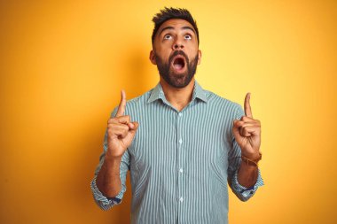 Yeşil çizgili tişört giyen genç Hintli adam izole edilmiş sarı arka planda duruyor. Şaşırmış ve şaşırmış bir şekilde yukarı bakıyor ve parmaklarıyla işaret ediyor ve kollarını kaldırıyor..