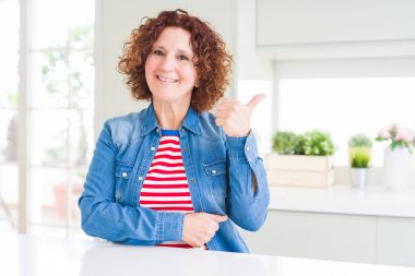 Orta yaşlı, kıvırcık saçlı, kot ceket giyen, gülümseyen, gülümseyen ve baş parmağıyla yüzünü gösteren bir kadın..