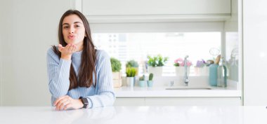 Bir öpücük el ile güzel ve seksi olmak hava üfleme kamera geniş açı resim güzel genç kadının evde beyaz masaya oturan bakarak. Aşk ifade.