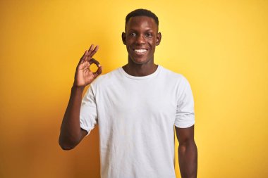 Beyaz t-shirt giyen genç Afro-Amerikalı adam izole edilmiş sarı arka planda duruyor. Gülümsüyor ve el ve parmaklarıyla 