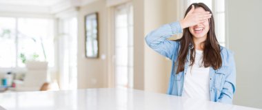 Güzel genç kadın masada gülümseyen ve ile Gülen gözler için sürpriz kapsayan yüz el beyaz evde oturan geniş açı resim. Kör kavramı.