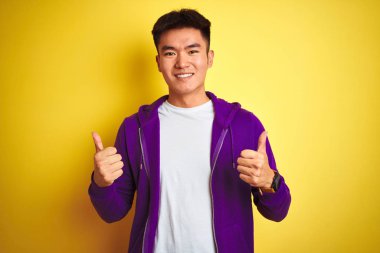 Mor kazak giyen Asyalı genç adam izole edilmiş sarı arka plan başarı tabelasının üzerinde duruyor. Elleriyle olumlu jest yapıyor, gülümsüyor ve mutlu. Neşeli ifade ve kazanan jest.