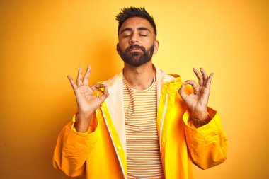 İzole edilmiş sarı arka planda duran yağmurluk giyen genç Hintli adam rahatla ve parmaklarıyla meditasyon hareketi yaparken gözleri kapalı gülümse. Yoga konsepti.