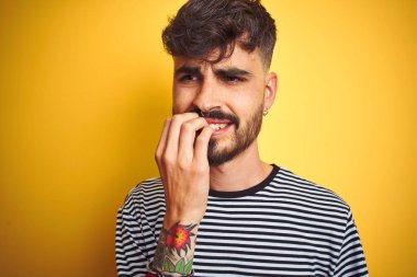 Çizgili t-shirt giyen dövme giyen genç adam izole sarı arka plan üzerinde duran stresli ve ağız tırnak ısırma elleri ile sinir görünümlü. Anksiyete sorunu.