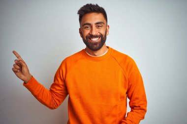 İzole edilmiş beyaz arka planda turuncu süveter giyen genç Hintli adam yüzünde kocaman bir gülümseme, eli ve parmağı kameraya bakıyor..