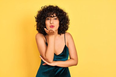 Sarı arka planda zarif elbiseler giyen kıvırcık saçlı genç bir Arap kadın havadaki güzel ve seksi elleriyle öpüşen kameraya bakıyor. Aşk ifadesi.