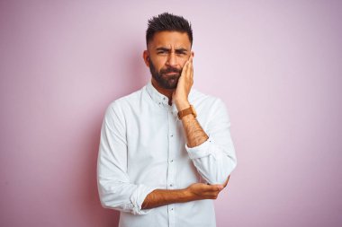 Genç Hintli iş adamı zarif bir gömlek giyip pembe arka planda duruyor ve kollarını kavuşturup depresyondan sıkılmış ve yorgun görünüyor..