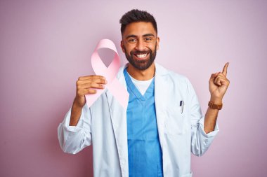 Kanser kurdelesi tutan genç Hintli doktor soyutlanmış pembe arka planda çok mutlu bir şekilde elini ve parmağını kenarı işaret ediyor.