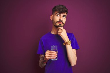 Dövmeli genç adam bir bardak su içiyor. Mor arka planda duruyor. Ciddi bir yüz.