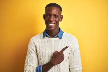İzole edilmiş sarı arka planda beyaz süveter ve kot gömlek giyen Afro-Amerikalı bir adam. Yüzünde mutlu ve doğal bir ifadeyle el ve parmağıyla işaret eden bir gülümseme var.