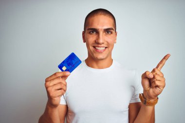 Genç ve yakışıklı bir adam kredi kartını soyutlanmış beyaz arka planda tutarken çok mutlu bir şekilde elini ve parmağını kenarı işaret ediyor.