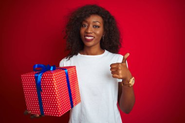 Genç Afrikalı Amerikalı bir kadın doğum günü hediyesi tutuyor. Kırmızı arka planda duruyor. Kocaman gülümsüyor.