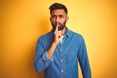 Kotlu tişört giyen genç Hintli adam izole edilmiş sarı arka planda duruyor ve dudakları parmaklarıyla sessiz olmak istiyor. Sessizlik ve gizli konsept.