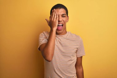 Çizgili tişört giyen genç yakışıklı Arap adam izole edilmiş sarı arka planda duruyor bir gözünü eliyle kapatıyor, yüzünde kendine güvenen bir gülümseme ve sürpriz bir duygu var..
