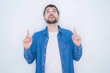 Beyaz yalıtılmış arka planda kot ceket giyen genç, yakışıklı İspanyol adam şaşırmış ve şaşırmıştı. Parmaklarıyla işaret ediyor ve kollarını kaldırıyordu..