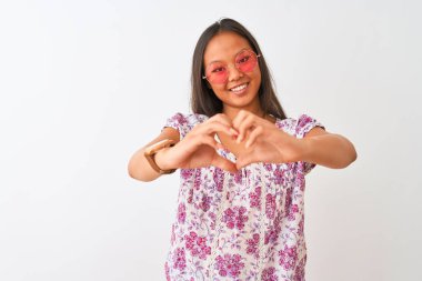 Çiçekli tişört ve pembe gözlük takan genç Çinli kadın izole edilmiş beyaz arka planda kalp sembolü ve ellerle şekil veren bir gülümsemeyle gülümsüyor. Romantik konsept.