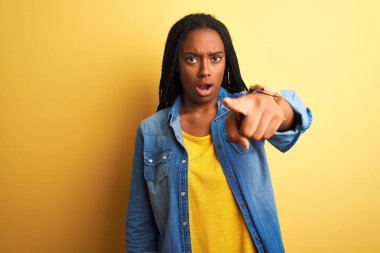 Genç Afro-Amerikalı kadın kot gömlek giyip izole edilmiş sarı arka planda dikiliyor. Kameraya kızgın ve sinirli olduğunu gösteriyor.