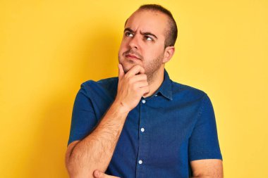 Kotlu tişört giyen genç bir adam izole edilmiş sarı arka planda duruyor ve elleri çenesinde soru sormayı düşünüyor, düşünceli bir ifade. Düşünceli bir yüzle gülümsüyor. Şüphe kavramı.
