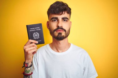Dövmeli, İtalyan pasaportlu genç bir adam izole edilmiş sarı bir arka planı var. Akıllı bir yüz ifadesi var ve ciddi düşünüyor.
