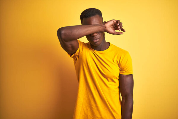 Young african american man wearing casual t-shirt standing over isolated yellow background covering eyes with arm, looking serious and sad. Sightless, hiding and rejection concept