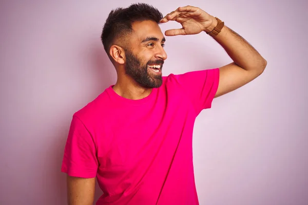 孤立したピンクの背景の上に立ってTシャツを着て若いインド人男性は非常に幸せと笑顔遠くに手を頭の上に見て立っている 検索の概念 — ストック写真