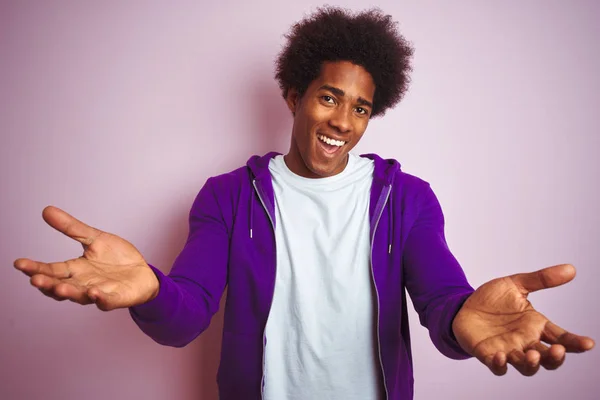 若いアフリカ系アメリカ人男性紫色のトレーナーを着て孤立したピンクの背景の上に立って笑顔明るく手を提供援助と受け入れを与える — ストック写真