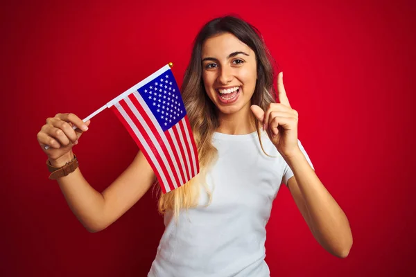 Young woman holding united states of america flag over red isolated background surprised with an idea or question pointing finger with happy face, number one