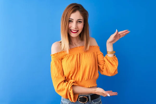 Beautiful redhead woman wearing orange casual t-shirt standing over isolated blue background gesturing with hands showing big and large size sign, measure symbol. Smiling looking at the camera. Measuring concept.
