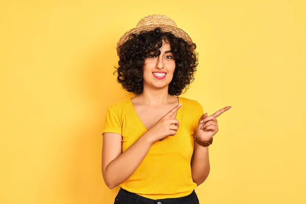 年轻的阿拉伯女人 卷曲的头发 穿着T恤衫 头戴黄褐色的帽子 面带微笑地看着摄像机 双手和手指指向旁边 — 图库照片