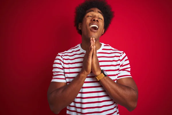 Young african american man with afro hair wearing striped t-shirt over isolated red background begging and praying with hands together with hope expression on face very emotional and worried. Asking for forgiveness. Religion concept.