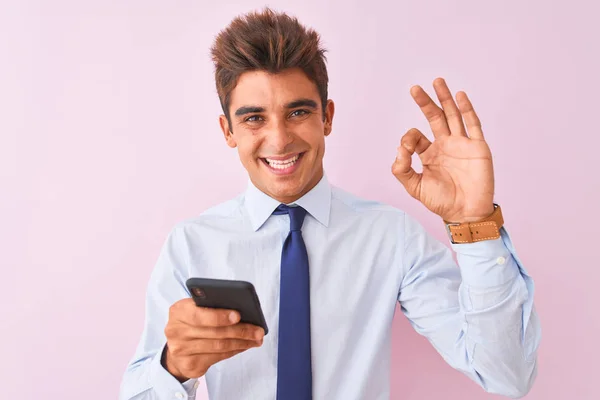 年轻英俊的商人用智能手机站在孤立的粉色背景上 手握手签 很好的符号 — 图库照片