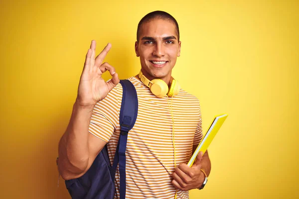 若いです学生男身に着けていますヘッドフォンとバックパック以上黄色隔離された背景Doing Signとともに指 優れたシンボル — ストック写真