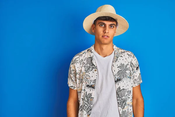 Indian man on vacation wearing hawiaian shirt summer hat over isolated blue background with serious expression on face. Simple and natural looking at the camera.