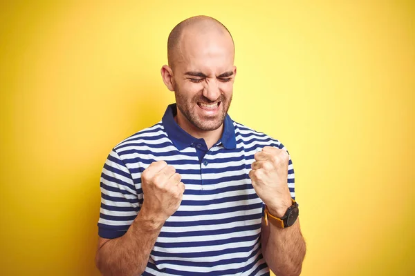 ひげを生やした若いはげ男は 黄色の孤立した背景にカジュアルな縞模様の青いTシャツを着て非常に幸せと興奮して腕を上げ 笑顔と成功のために叫んで勝者のジェスチャーをしています お祝いのコンセプト — ストック写真