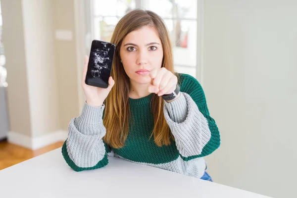 Beautiful young woman holding broken smartphone pointing with finger to the camera and to you, hand sign, positive and confident gesture from the front