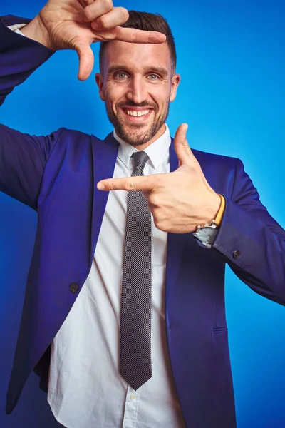 Vertical angle picture of young handsome business man over blue isolated background smiling making frame with hands and fingers with happy face. Creativity and photography concept.