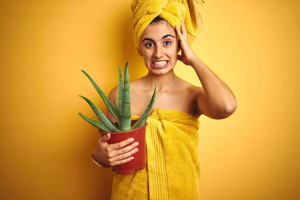 Young beautiful woman wearing a towel holding cactus pot over yellow isolated background stressed with hand on head, shocked with shame and surprise face, angry and frustrated. Fear and upset for mistake.