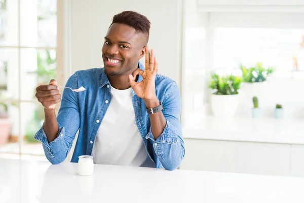 African american man eating healthy natural yogurt with a spoon doing ok sign with fingers, excellent symbol
