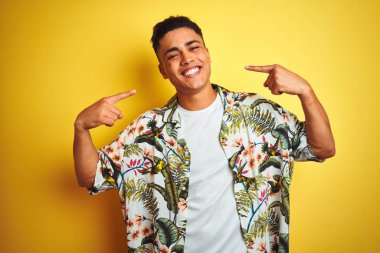 Tatilde olan genç Brezilyalı bir adam izole edilmiş sarı arka plan üzerinde çiçekli gömlek giyiyor neşeli bir şekilde gülümsüyor ve parmakları ve ağzı ile işaret ediyor. Diş sağlığı konsepti.