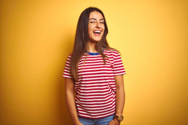 Çizgili tişört giyen genç ve güzel bir kadın izole edilmiş sarı arka planda duruyor kameraya seksi bir ifadeyle bakıyor, neşeli ve mutlu bir yüzle..